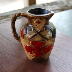 May include: A small, decorative ceramic pitcher with a brown base color and a dark brown interior. The pitcher features a braided handle and is adorned with red, green, and black painted designs, including crossed axes and a stylized bird.