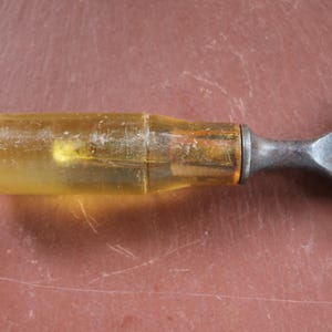 Puede incluir: Primer plano de un cincel con un mango transparente teñido de amarillo y una hoja de metal oscuro. El mango parece estar hecho de plástico translúcido. El cincel está sobre una superficie marrón rojiza.