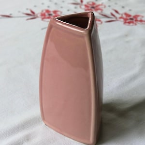 May include: A dusty rose-colored ceramic vase with a triangular opening and a rectangular body. The vase has a smooth, glossy finish and a slightly curved shape. The vase is approximately 8 inches tall.