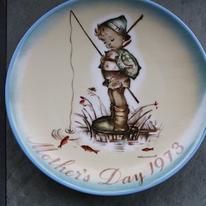 Può includere: Un piatto in ceramica vintage con un bordo blu presenta un dipinto di un giovane ragazzo che pesca. Il ragazzo indossa un cappello verde e stivali marroni. Il piatto ha il testo "Mother's Day 1973" sul fondo.