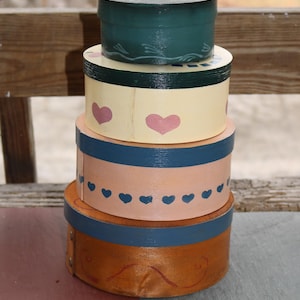 May include: A stack of four round wooden boxes with painted designs. The top box is green with white accents, the second box is yellow with green accents and pink hearts, the third box is pink with blue accents and small blue hearts, and the bottom box is brown with blue accents and a swirl design.