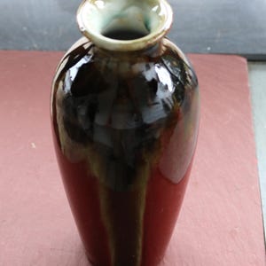 May include: A handcrafted ceramic vase with a glossy finish. The vase features a color gradient, transitioning from a deep red base to a dark brown midsection, and a light green rim. The vase has a smooth, rounded shape.