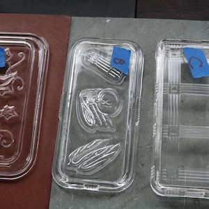 May include: Three clear glass rectangular dishes with embossed designs. One features a floral pattern, another has a fly and asparagus design, and the third has a grid pattern. Each dish has a blue tape label.