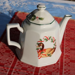 May include: White porcelain teapot with a green trim and a festive design featuring a duck, holly, and red ribbons. The teapot has a rounded body and a lid with a knob.
