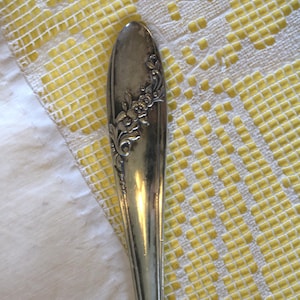 Könnte beinhalten: Nahaufnahme eines Vintage-Silberlöffels mit floralen Details. Der Löffelstiel weist ein geprägtes Blumendesign auf. Der Löffel befindet sich vor einem gelb-weiß gemusterten Hintergrund, was auf antikes Silberbesteck oder Vintage-Küchenutensilien hindeutet.