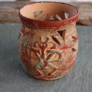 Puede incluir: Un portavelas de cerámica hecho a mano con un acabado rústico y texturizado. El recipiente cilíndrico presenta diseños recortados de hojas y geométricos, pintados en tonos rojos, verdes y beige. El borde está pintado de rojo.