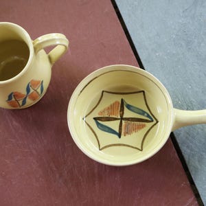 May include: A pale yellow ceramic creamer and a matching small handled bowl. Both pieces feature a hand-painted geometric design in orange, blue, and dark brown. The creamer has a pouring spout and a side handle, while the bowl has a single handle.
