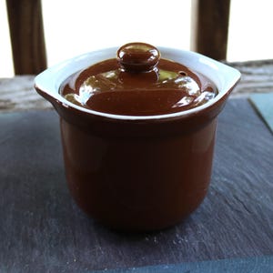 Może przedstawiać: Małe, brązowe ceramiczne naczynie do zapiekania z pokrywką. Naczynie ma białą obwódkę i mały, okrągły uchwyt na pokrywce. Naczynie do zapiekania stoi na ciemnoszarej powierzchni.