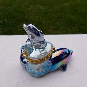 May include: A blue and iridescent porcelain trinket box shaped like a dolphin. The dolphin has a smaller dolphin on its back. The box has a gold hinge and a gold trim around the lid.