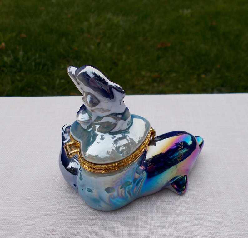 May include: A blue and iridescent porcelain trinket box shaped like a dolphin. The dolphin has a smaller dolphin on its back. The box has a gold hinge and a gold trim around the lid.