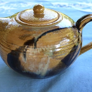 Puede incluir: Una tetera de cerámica con tapa y asa. La tetera está esmaltada en tonos marrones y beige con detalles en marrón oscuro y negro. La tetera está sobre una superficie azul.