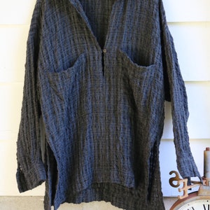 May include: A dark gray, textured, long-sleeved shirt with a button-up front and two large pockets. The shirt has a relaxed fit and a slightly longer hem in the back.