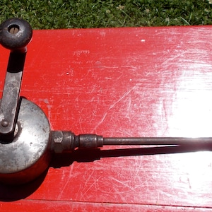 May include: A vintage hand-cranked drill with a metal body and a black wooden handle. The drill has a long, thin metal shaft extending from the side.