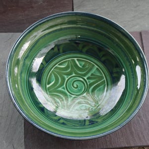 May include: A handmade ceramic bowl with a glossy green glaze. The bowl features a spiral design in the center and a dark blue rim. The bowl is round and has a wide opening, perfect for serving salads or displaying as decor.