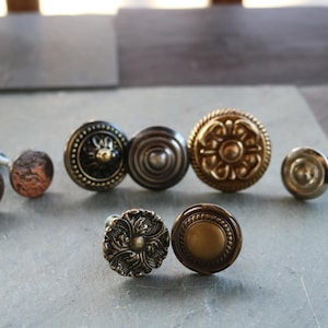May include: A collection of antique cabinet knobs in various styles and finishes. The knobs include round, textured, and ornate designs in bronze, brass, and silver tones. Some knobs have a hammered appearance, while others feature intricate floral patterns.