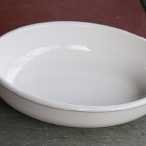 Puede incluir: Una fuente de horno ovalada de cerámica blanca con dos asas. La fuente tiene un acabado liso y brillante y está diseñada para usar en el horno. La fuente está vacía y se encuentra sobre una superficie oscura.