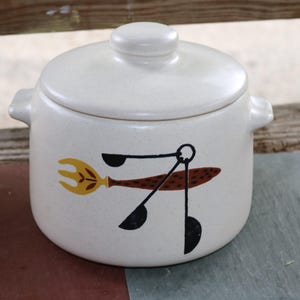 Peut inclure: Casserole en céramique crème avec couvercle. Le plat présente un graphisme de fourchette, de cuillère et de spatule en jaune, marron et noir. Il possède deux poignées latérales et un couvercle rond avec un petit bouton.