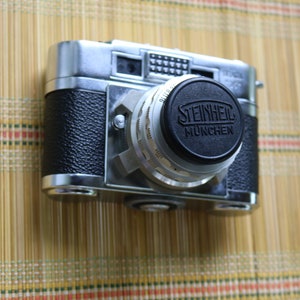 Pu&ograve; includere: Una macchina fotografica a pellicola da 35 mm argentata e nera con un tappo dell'obiettivo che dice "STEINHEIL M&Uuml;NCHEN".