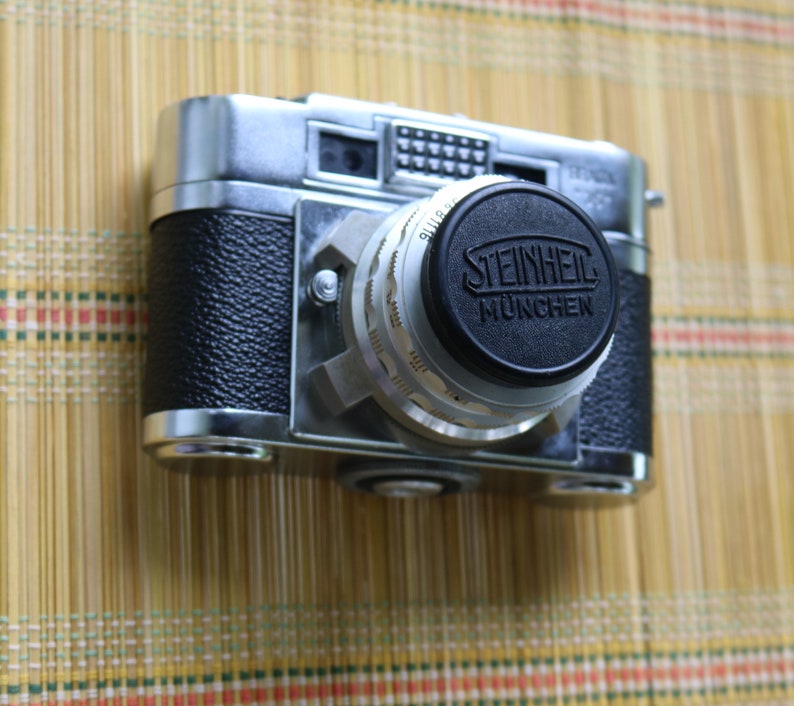 Pu&ograve; includere: Una macchina fotografica a pellicola da 35 mm argentata e nera con un tappo dell'obiettivo che dice "STEINHEIL M&Uuml;NCHEN".