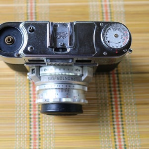 Pu&ograve; includere: Una macchina fotografica a pellicola da 35 mm di colore argento con un obiettivo nero e un esposimetro sulla parte superiore. La macchina fotografica &egrave; etichettata "PRONTOR-SVS".