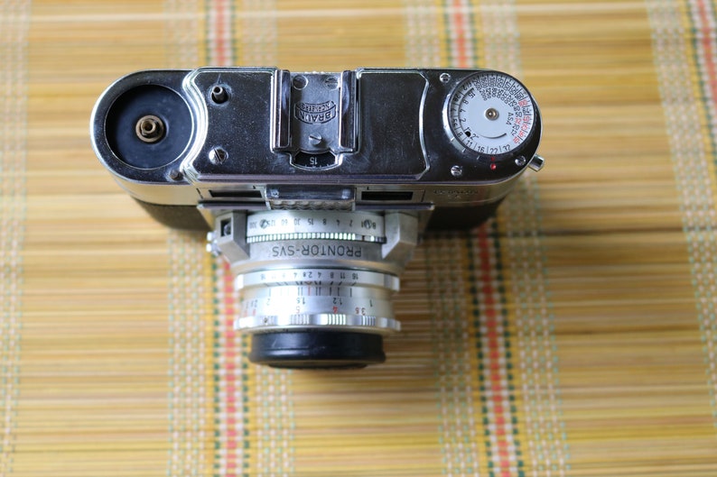 Pu&ograve; includere: Una macchina fotografica a pellicola da 35 mm di colore argento con un obiettivo nero e un esposimetro sulla parte superiore. La macchina fotografica &egrave; etichettata "PRONTOR-SVS".