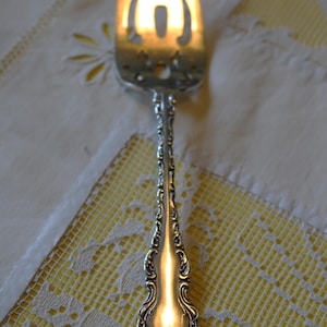 Pode incluir: Close-up de um garfo de servir prateado com um cabo ornamentado e uma cabeça decorativa. O garfo está sobre uma toalha de mesa de renda branca e amarela. O design do garfo é realçado pela luz.
