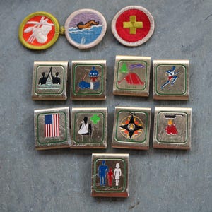 May include: A selection of vintage Scout badges and metal clips. The badges show embroidered designs of a campfire, a swimmer, and a first aid cross. The metal clips depict activities such as camping and first aid, with various illustrations.