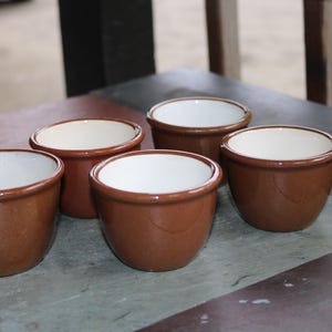 May include: Five small, brown ceramic bowls with a glossy finish and white interiors are arranged on a gray surface. The bowls have a rounded shape and a simple, classic design, suitable for serving dips or small portions.