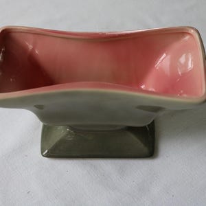 May include: A vintage ceramic planter with a rectangular shape and a two-tone color scheme. The interior is a soft pink, while the exterior and base are a muted green. The planter has a curved, open top.
