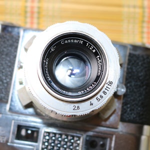 Pu&ograve; includere: Un obiettivo per fotocamera argentato con il testo "Cassarit 1:2.8 f=50mm" e "Steinheil M&uuml;nchen" stampato sul lato. L'obiettivo ha un anello di apertura nero con i numeri 2.8, 4, 5.6, 8, 11 e 16 stampati su di esso.
