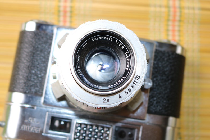 Pu&ograve; includere: Un obiettivo per fotocamera argentato con il testo "Cassarit 1:2.8 f=50mm" e "Steinheil M&uuml;nchen" stampato sul lato. L'obiettivo ha un anello di apertura nero con i numeri 2.8, 4, 5.6, 8, 11 e 16 stampati su di esso.