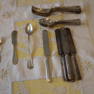 Può includere: Un assortimento di posate antiche, tra cui forchette, cucchiai, coltelli da burro e coltelli da tavola, disposte su una tovaglia bianca con ricami floreali gialli. Le posate hanno una finitura metallica lucida.