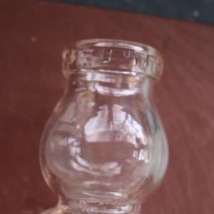 May include: A clear glass bottle with a rounded body and a narrow neck. The bottle has a small, textured rim at the top. The glass is transparent, allowing a view of the interior. The bottle is likely for storing liquids.