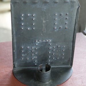 May include: A dark gray metal wall sconce featuring a house-shaped backplate with a series of small punched holes arranged in patterns. The base includes a cylindrical candle holder with a vertical slit, and a small hole at the top for mounting.