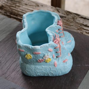 Könnte beinhalten: Hellblauer Keramik-Pflanztopf in Stiefelform mit rosa und gelben floralen Akzenten. Der Pflanztopf hat rosa Schnürsenkel und ein spitzenartiges Muster auf der Vorderseite. Der Innenraum ist einfarbig hellblau.