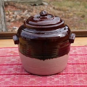 Op de afbeelding: Een bruine en roze geglazuurde keramische pot met een deksel. De pot heeft een rond lichaam en twee handvatten. Het deksel heeft een knop bovenop.