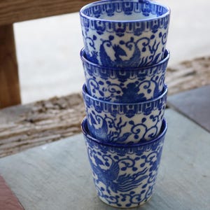 May include: Stack of four blue and white ceramic teacups with a phoenix design. The cups are stacked on a wooden surface.
