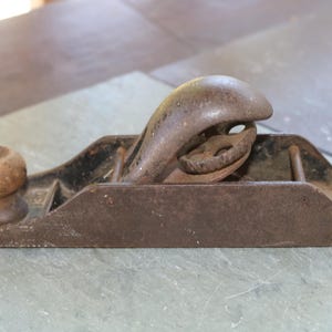以下が含まれることがあります： 使い込まれた茶色の仕上げが施されたヴィンテージの金属製ハンドプレーン。丸みを帯びた木製のフロントノブと中央の調整ホイールが特徴です。経年変化と酸化の跡が見られる、低プロファイルの本体を持つクラシックなデザインの工具です。