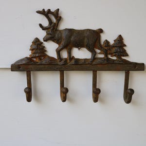 Puede incluir: Un perchero de pared rústico de metal con un diseño de ciervo y pino. El gancho marrón tiene cuatro ganchos para colgar objetos. La pieza decorativa está montada en una pared blanca.