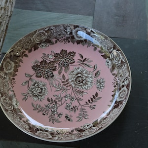 以下が含まれることがあります： 丸いピンク色のセラミックプレートで、花柄のデザインです。プレートの中央には、茶色と白の色合いの、大きく様式化された花と葉のパターンがあります。縁には繰り返しの花のボーダーがあります。プレートは暗い表面に置かれています。