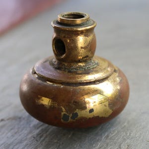 Może przedstawiać: Antyczny mosiężny nóżka meblowa z zaokrągloną podstawą i cylindrycznym wierzchem. Mosiądz ma ciepłą, postarzaną patynę z obszarami zużycia, odsłaniając metal. Nóżka ma mały otwór z boku i gwintowany otwór na górze.