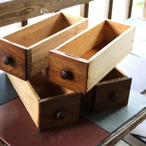 Op de afbeelding: Drie rechthoekige houten lades met een interieur van natuurlijk licht hout en donkerbruin gebeitste voorpanelen. Elke lade is voorzien van een centrale donkerbruine houten knop. De lades zijn in een overlappende opstelling gestapeld.