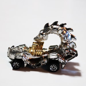 Puede incluir: Un coche de juguete plateado con forma de dragón con detalles dorados. El coche tiene un diseño detallado y es un juguete de colección.