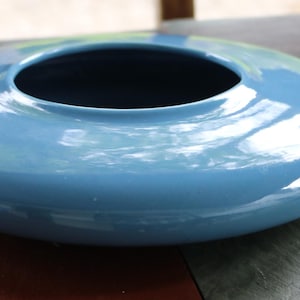 May include: A light blue ceramic bowl with a glossy finish. The bowl has a wide, oval shape with a circular opening in the center. The interior is dark. The bowl is likely used for decorative purposes.