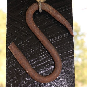 Puede incluir: Un gancho de metal oxidado con un gancho de latón en la parte superior, montado en una superficie de madera negra.