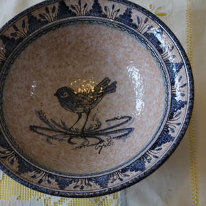 May include: Ceramic bowl with a bird illustration. The bowl has a beige interior with a blue bird design and a blue floral border. The bowl is round and has a glossy finish.