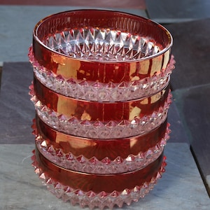 Può includere: Una pila di quattro ciotole di vetro rosso rubino. Ogni ciotola ha un interno trasparente tagliato a diamante e un esterno rosso, simile al metallo. I bordi e le basi presentano un design decorativo a punta. Le ciotole sono impilate su una superficie grigia.
