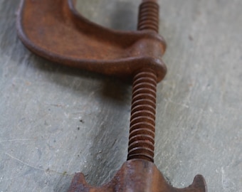May include: Close-up of a rusty C-clamp, a tool used for holding workpieces together. The clamp is made of metal and has a threaded screw for adjusting the pressure. The C-clamp is a reddish-brown color and shows signs of age and use.