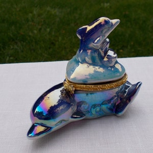 May include: A blue and iridescent porcelain trinket box shaped like a dolphin. The lid features a smaller dolphin figurine.