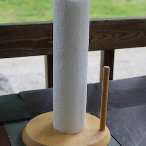 Könnte beinhalten: Ein hellfarbener Papierrollenhalter aus Holz mit rundem Fuß, einem zentralen vertikalen Stab und einem kleineren Seitenstab. Eine Rolle weißes Küchenpapier mit strukturiertem Muster steckt auf dem Mittelstab, gekrönt von einem runden Holzknauf.